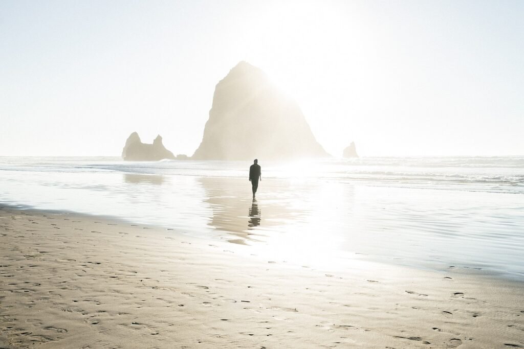 Person walking alone on a sunlit beach toward a large sea stack rock formation, with reflections on the wet sand.