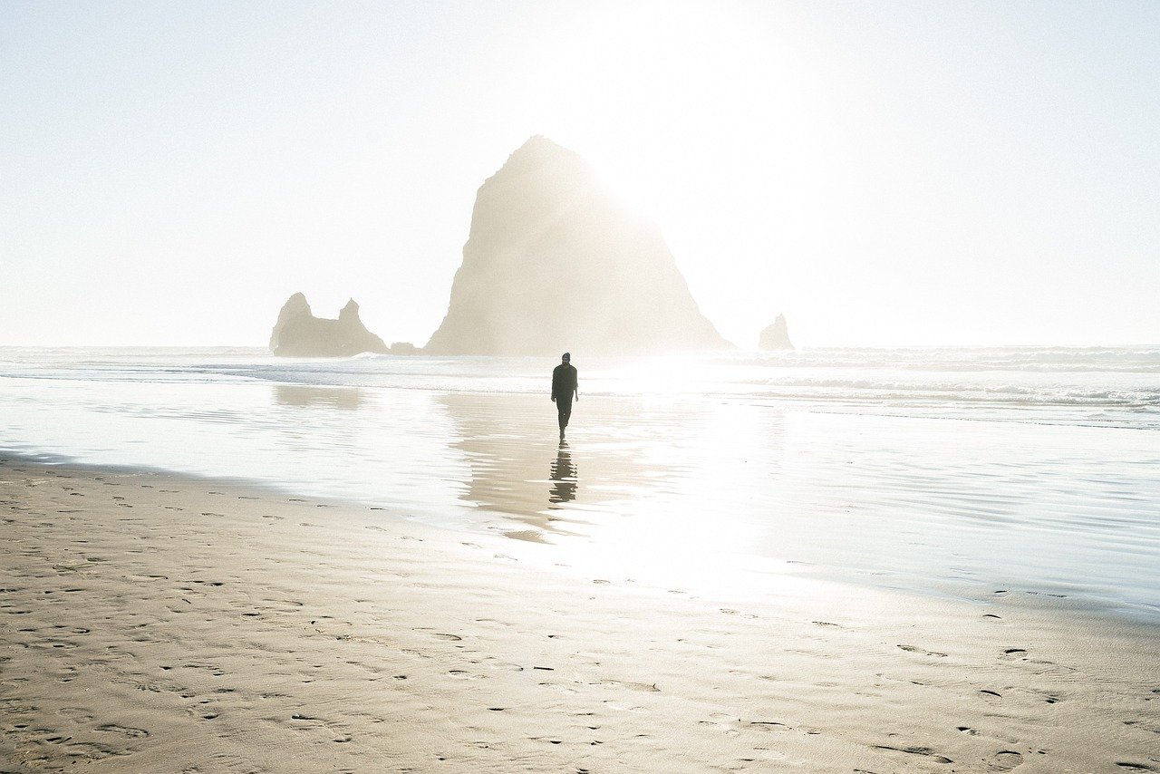 Men's Healing Journey- Person walking alone on a sunlit beach toward a large sea stack rock formation, with reflections on the wet sand.